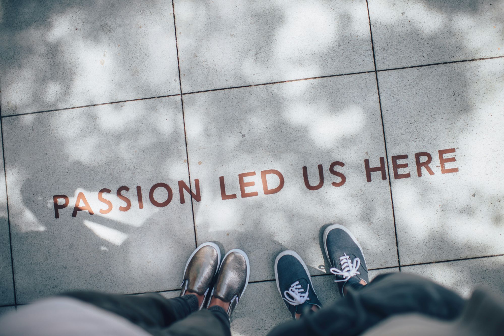 Two people's feet standing next to a sign on the ground that says "passion led us here"
